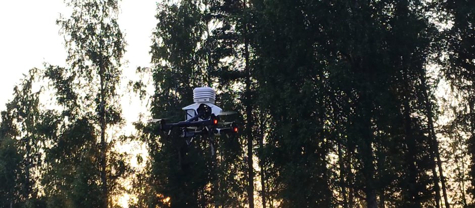 A WeatherSens drone hovering at treetop height against a low sun, its meteorological sensor shield on top.